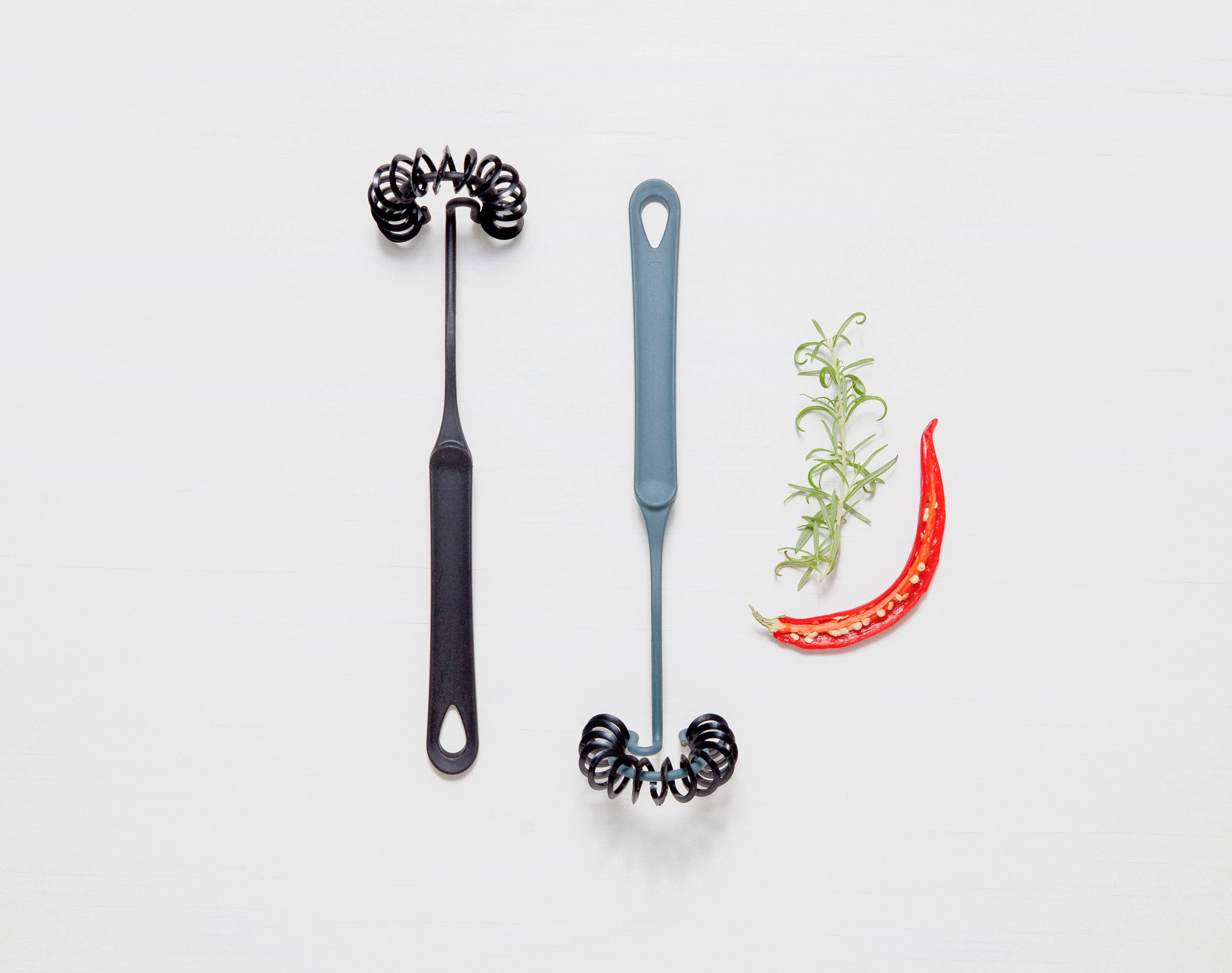 Two black Gourmaid Whisks with spiral ends, made from BPA-free, heat-resistant silicone, are placed beside sliced red chili and fresh rosemary on a white background.