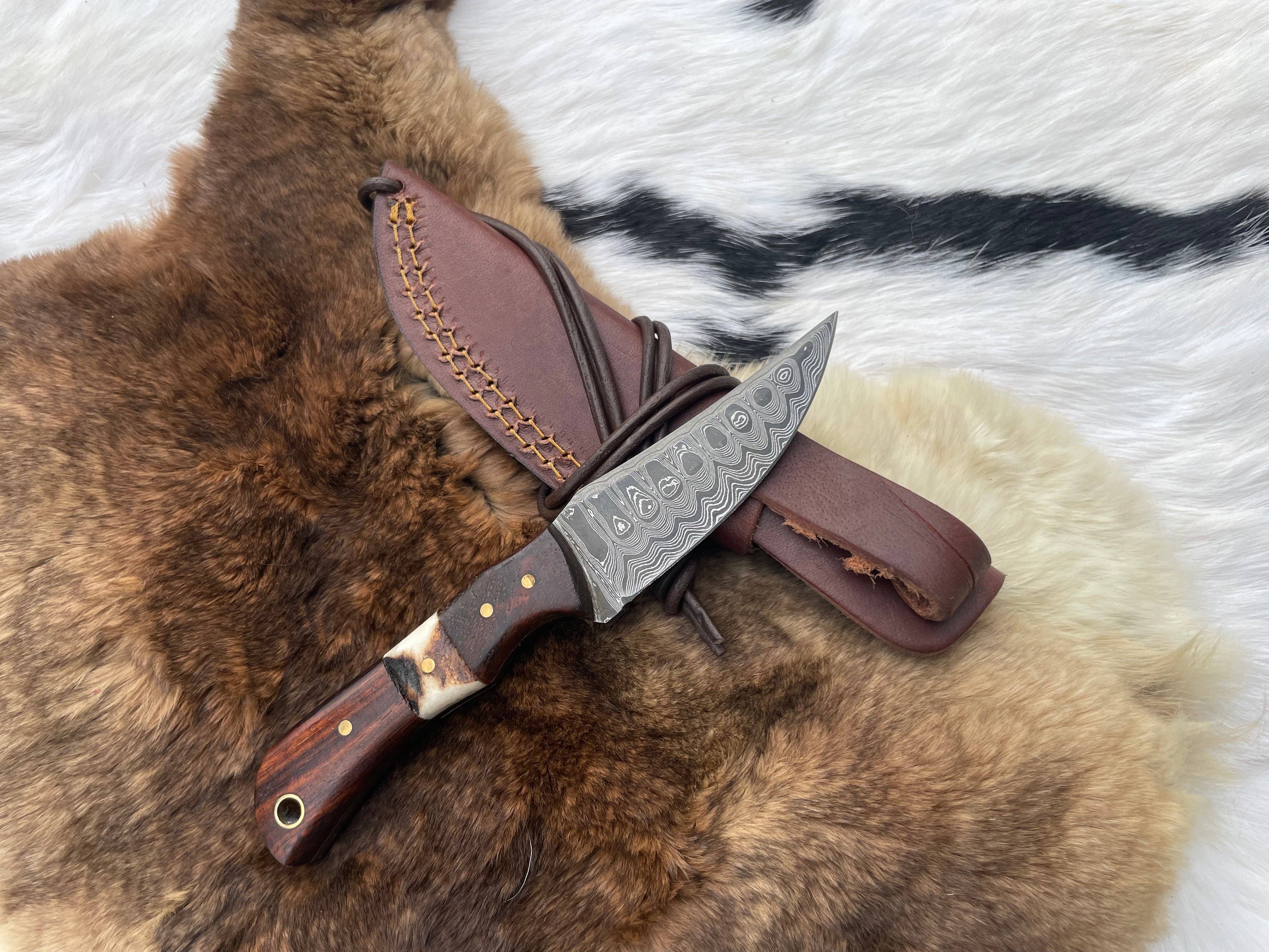 The Knife: Damascus Steel Skinner Knife with Rosewood & Stag Grip lies on a fur surface next to its brown leather sheath.