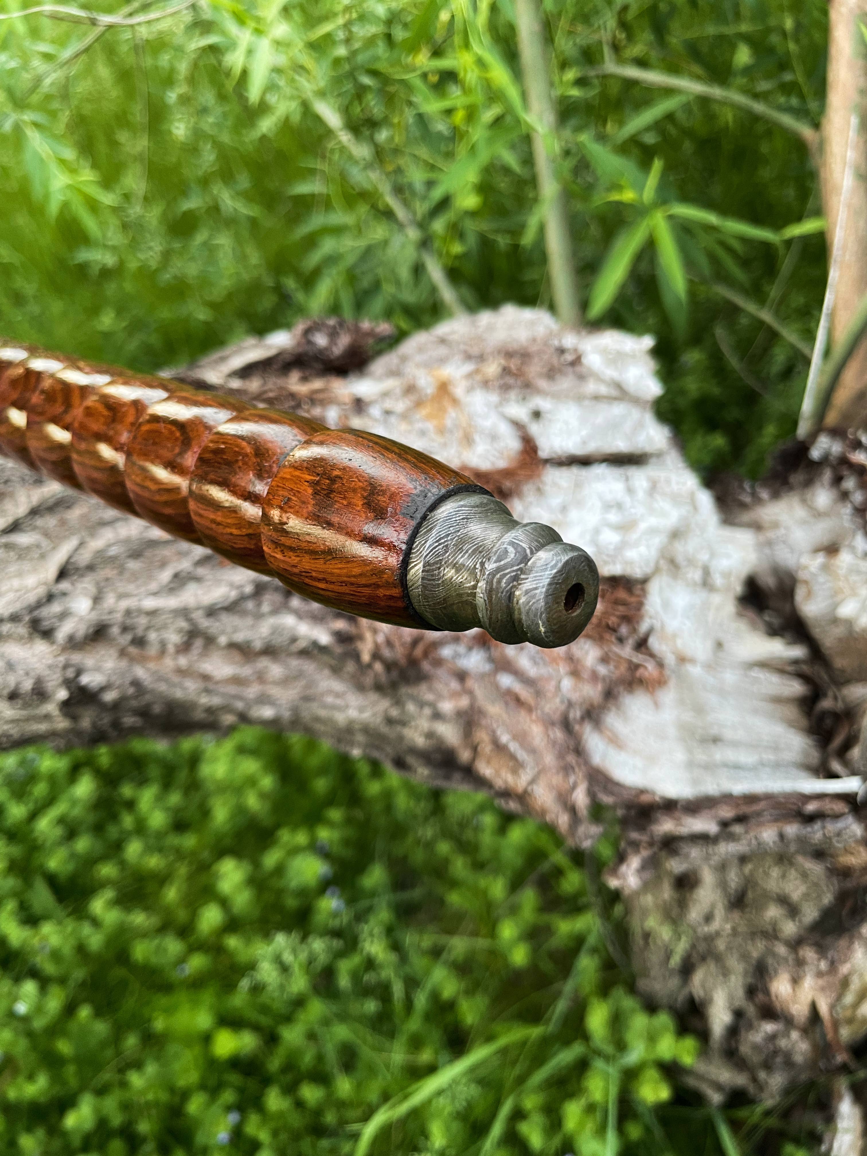 Close-up of the carved wooden handle and metal tip of the Axe: Peace Pipe Axe Handmade with Damascus Steel, set against green foliage and a fallen tree in the background.