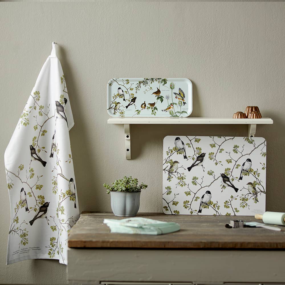 A kitchen scene with a tea towel, placemat, and the Garden Birds birch veneer serving tray (14.9 diameter) by Koustrupco of Denmark—all with bird and branch illustrations—displayed on a wooden counter and shelf.