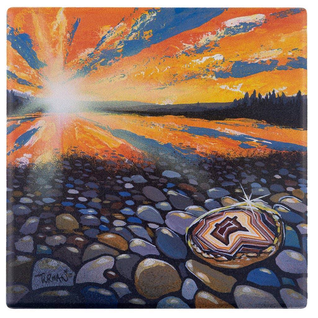 A bright sunset over a lake reflects vivid orange and blue hues. In the foreground, rocks line the shore alongside a Coaster: Discover Agates - Minnesota 4x4 in Absorbent Stone Coaster with Cork Back by Adam Turman sparkling in the sunlight.