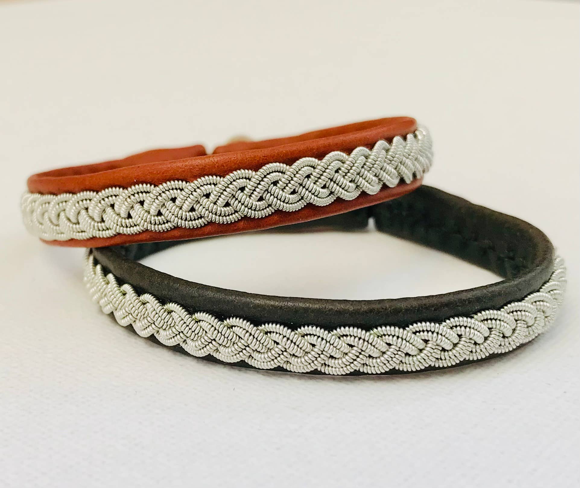 A dark brown Lapland Sami bracelet crafted from reindeer hide with a single pewter and silver braid is displayed on a white surface.