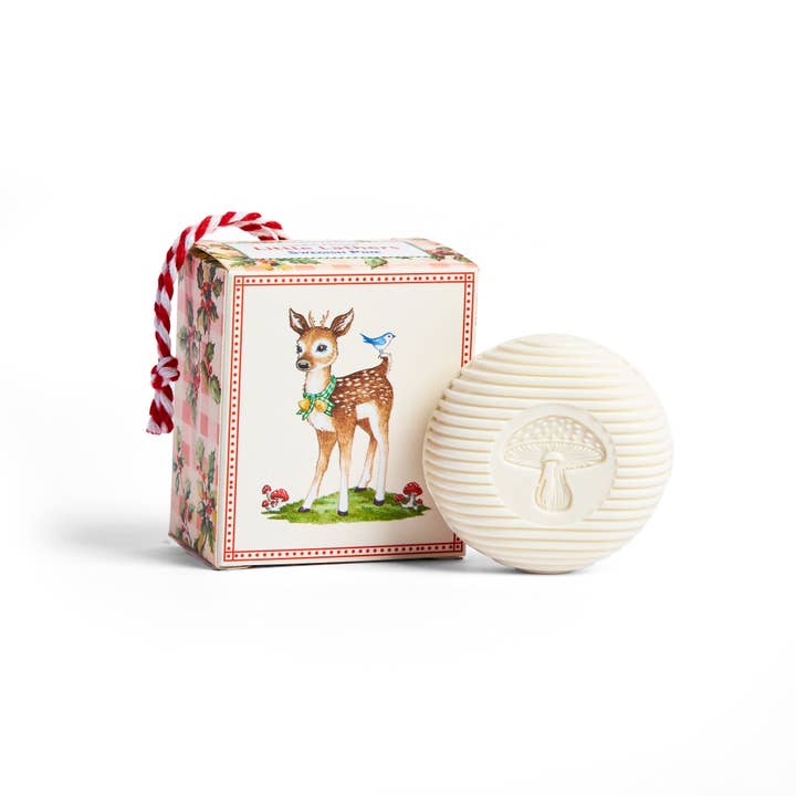 A round, ivory Swedish Dream Little Lathers Swedish Pine bar soap embossed with a mushroom rests beside a decorative box with a fawn, bluebird, and a red-white rope handle—adding a whimsical holiday touch.