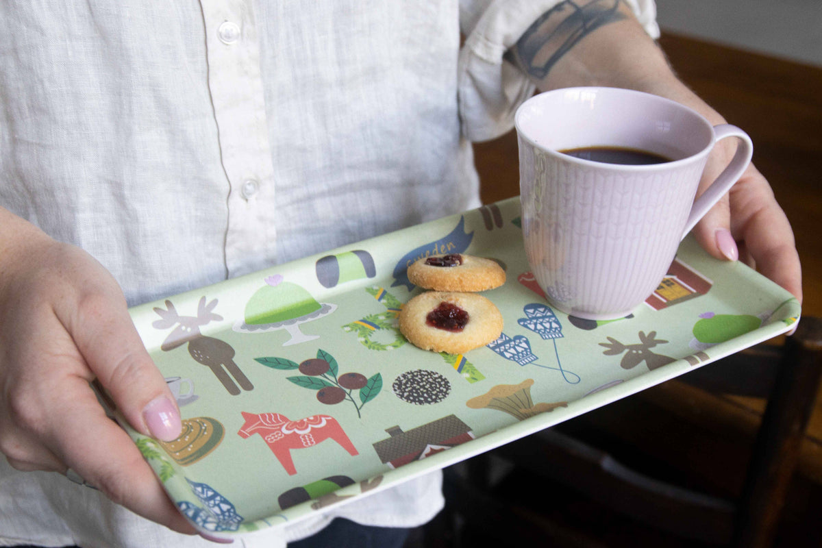 Person holding a bamboo tray featuring Dala horse and winter patterns, two jam-centered cookies, and a pale pink mug of coffee.