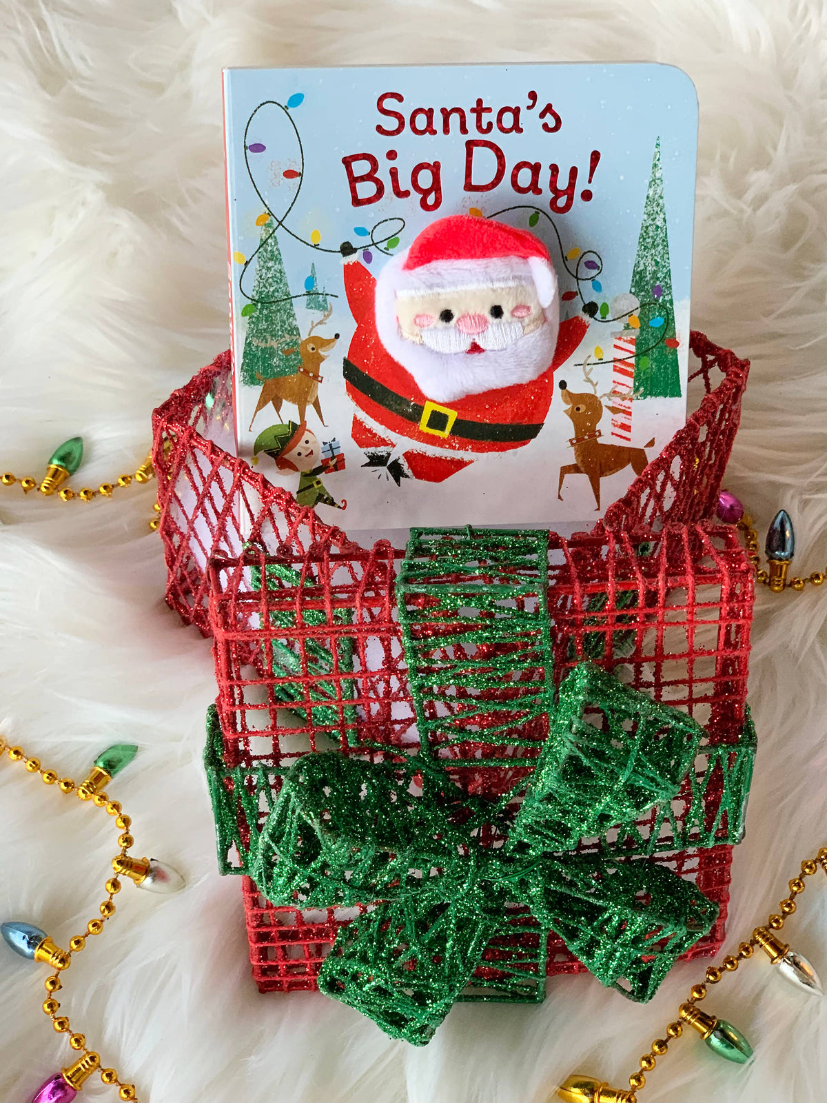 A board book with a Santa finger puppet topper, positioned inside a red and green mesh gift box ornament.