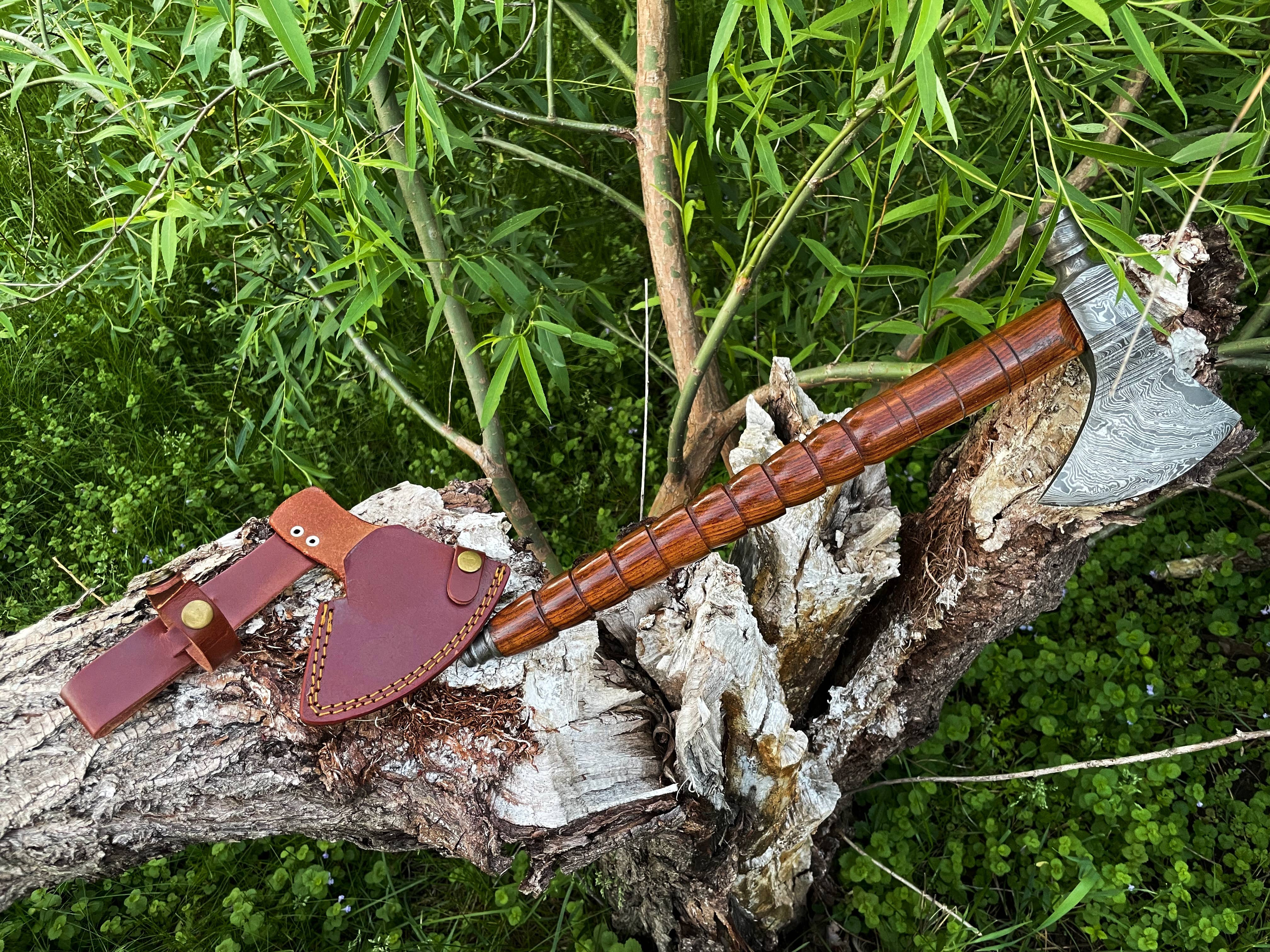 The Peace Pipe Axe Handmade with Damascus Steel rests on a fallen tree trunk, its custom sheath beside it, surrounded by green foliage and branches.