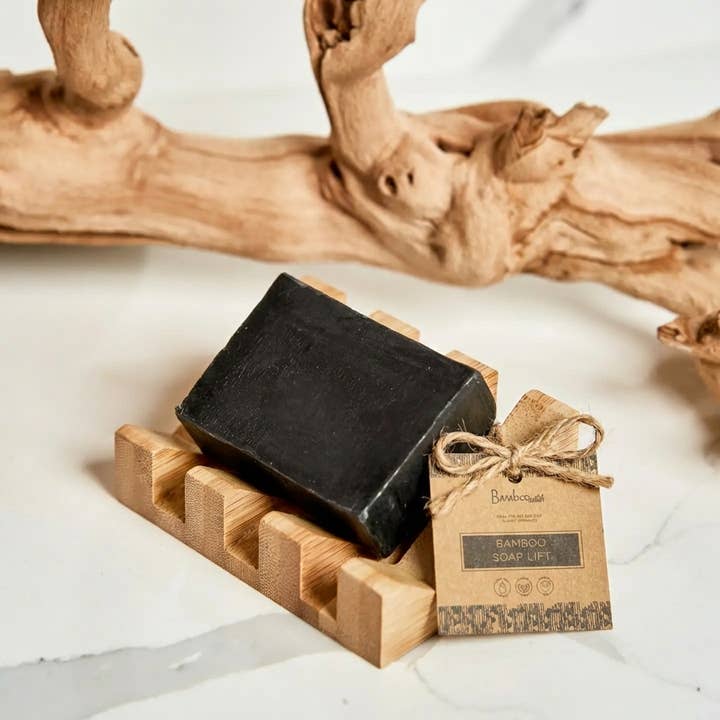 Black soap bar on a bamboo holder with a brown label tag tied in twine; large textured driftwood piece in the background.