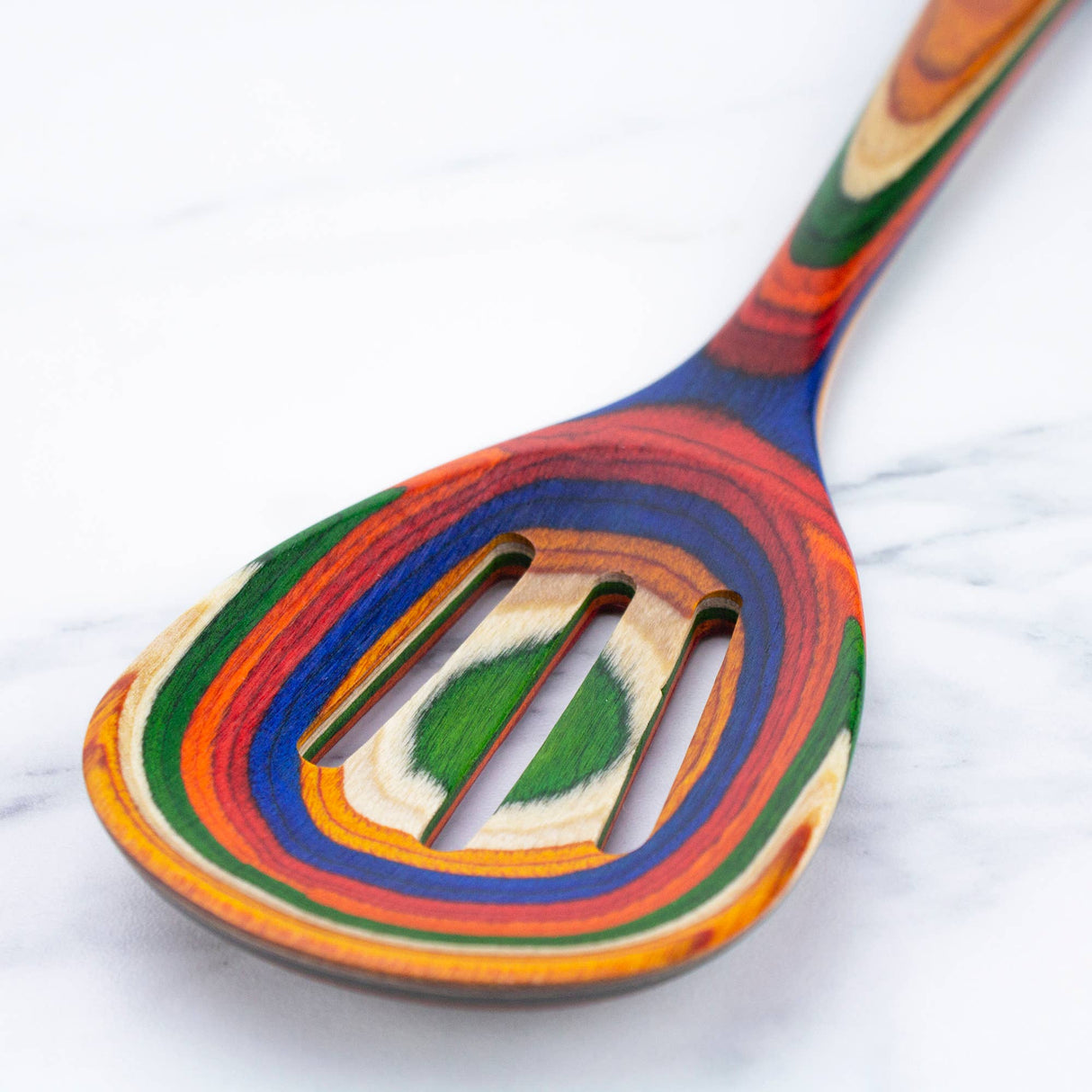 A close-up of the Marrakesh Collection Slotted Wooden Cooking Spoon displays its vibrant multicolored stripes and non-stick safe design as it rests elegantly on a white marble surface.