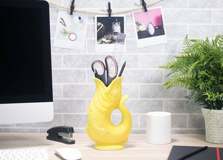 A yellow Gluggle Jug: Fish Shaped Pitcher that Gluggles, handmade in the UK, holds scissors and a ruler on a desk next to a computer, stapler, mug, plant, and hanging photos.