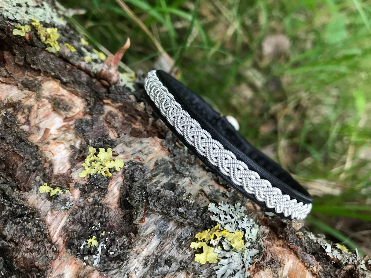 The Traditional Sami Single Braid Bracelet - Black #1, handcrafted in Lapland, Sweden from reindeer leather with a silver braid, rests on tree bark amid green grass and yellow lichen.