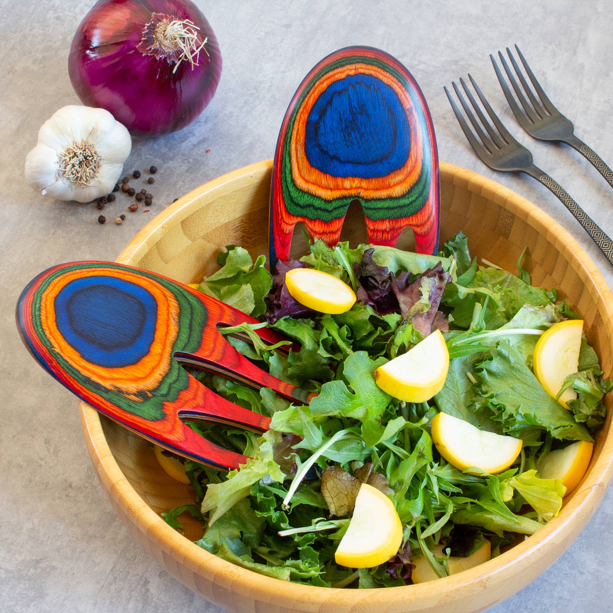 A wooden bowl of mixed green salad with yellow squash, Colorful Birch Salad Hands serving utensils, two forks, red onion, garlic, and peppercorns on a gray surface.