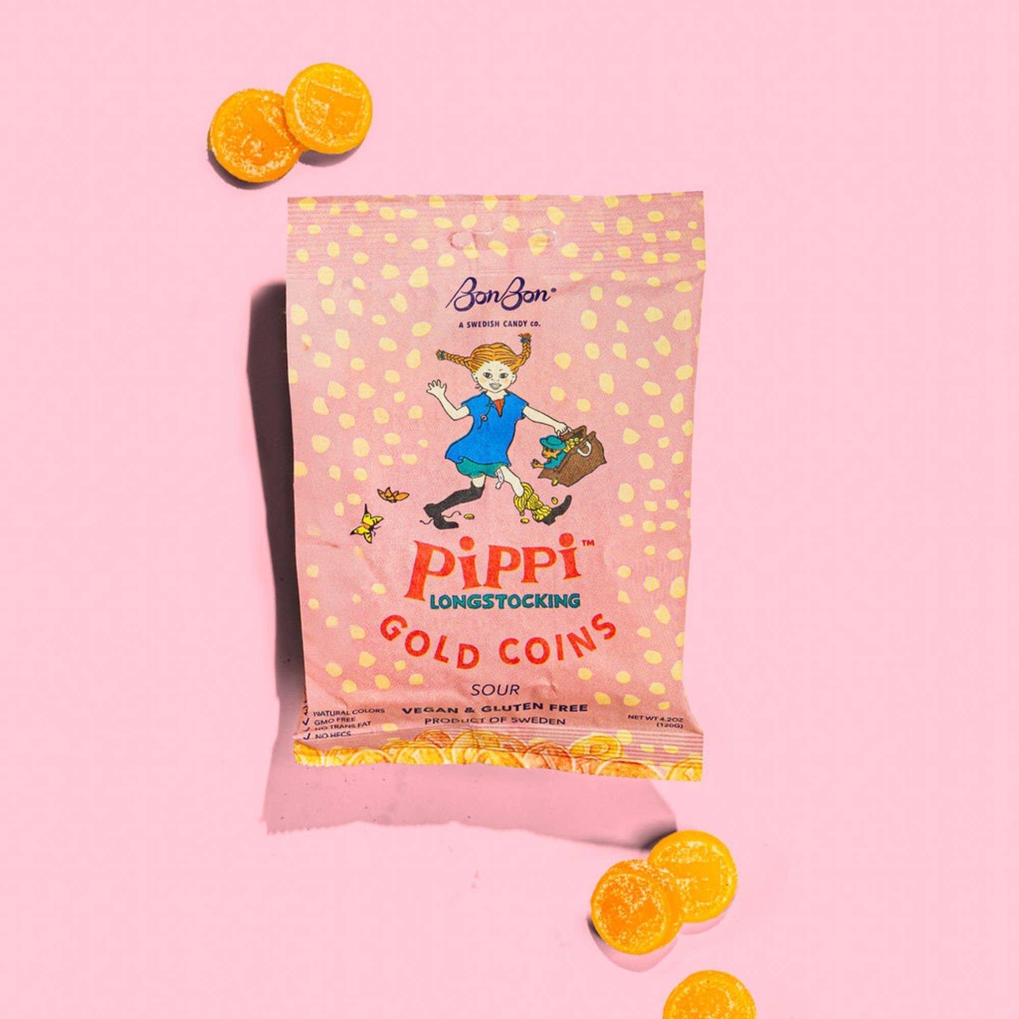 A pink bag labeled Candy: Pippi Longstocking Gold Coins - Swedish Candy 4.2oz (120g) with playful art is surrounded by yellow, coin-shaped tropical-flavored candies on a pink background.