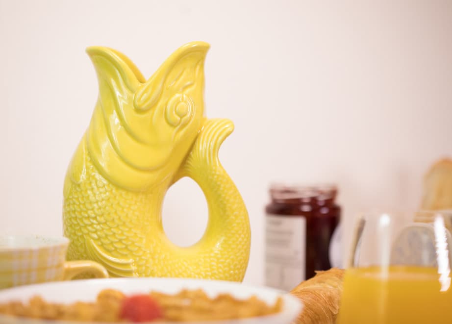 A Yellow Gluggle Jug: Fish Shaped Pitcher that Gluggles, handmade in the UK, sits on a breakfast table with cereal, orange juice, a croissant, and jam jar in the background.