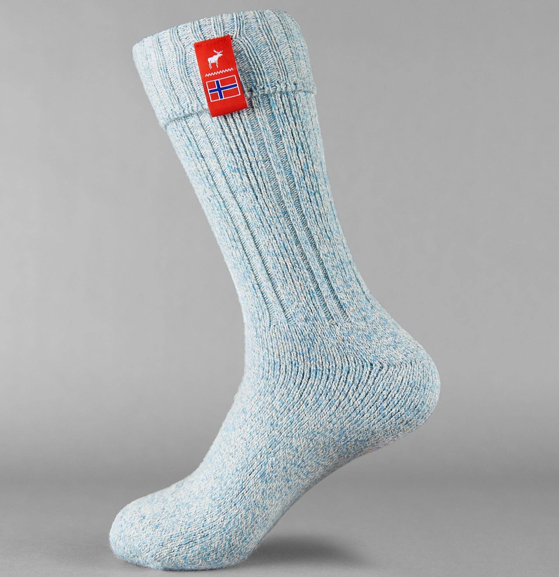 The Nordic Sock Company - Norwegian Fjord Socks in light blue, shown on a mannequin foot with a red tag featuring a white animal, and a blue-red-white flag patch—ideal for cozy comfort and perfect as wellie socks.