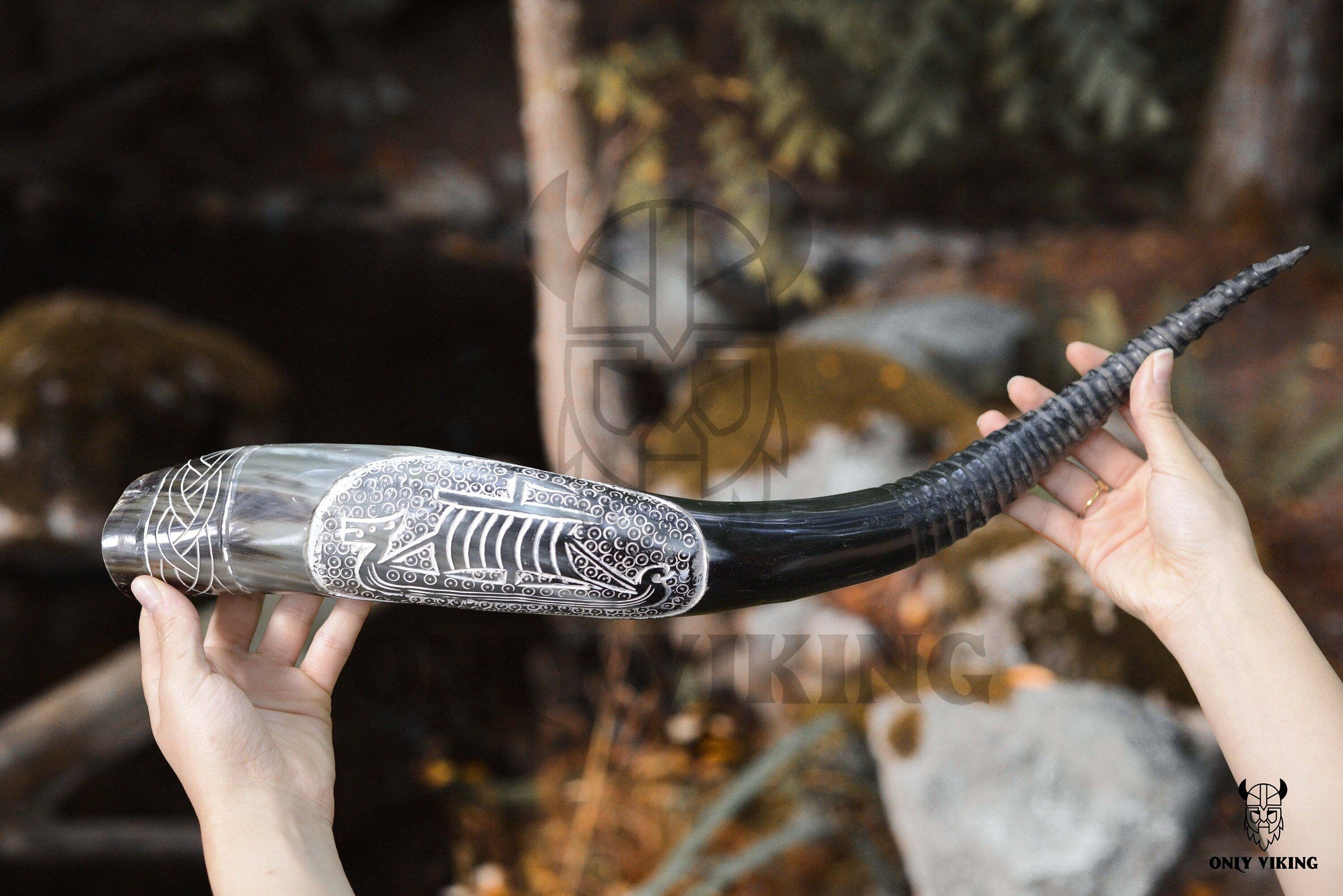 A person holds the Ravens Viking Ship Handcrafted Drinking Horn (14-18 inches) with intricate designs outdoors, while its metal stand rests nearby.