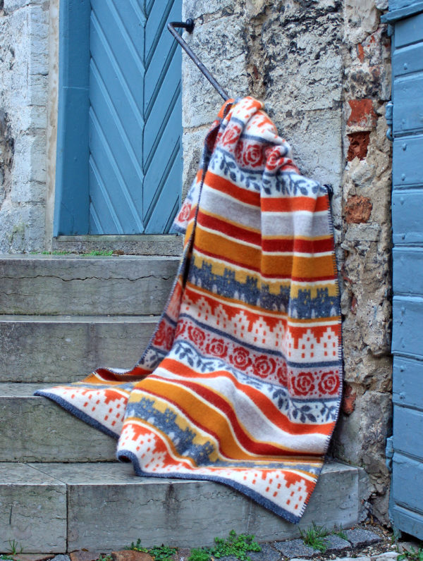 The Visby blanket in rust, designed by Kerstin Landström and made from OEKO-TEX certified wool, is elegantly draped over stone steps near a blue wooden door.