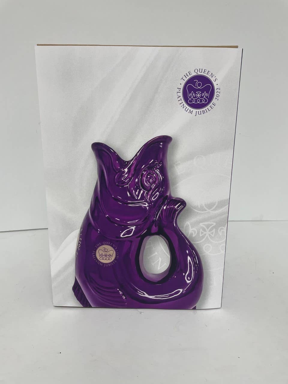 The Gluggle Jug: Limited Edition Queens Jubilee Gluggle Jug Extra Large, a purple ceramic fish-shaped jug, is displayed in front of a box marked with The Queen’s Platinum Jubilee 2022 emblem and seal—a unique commemorative collectors piece.