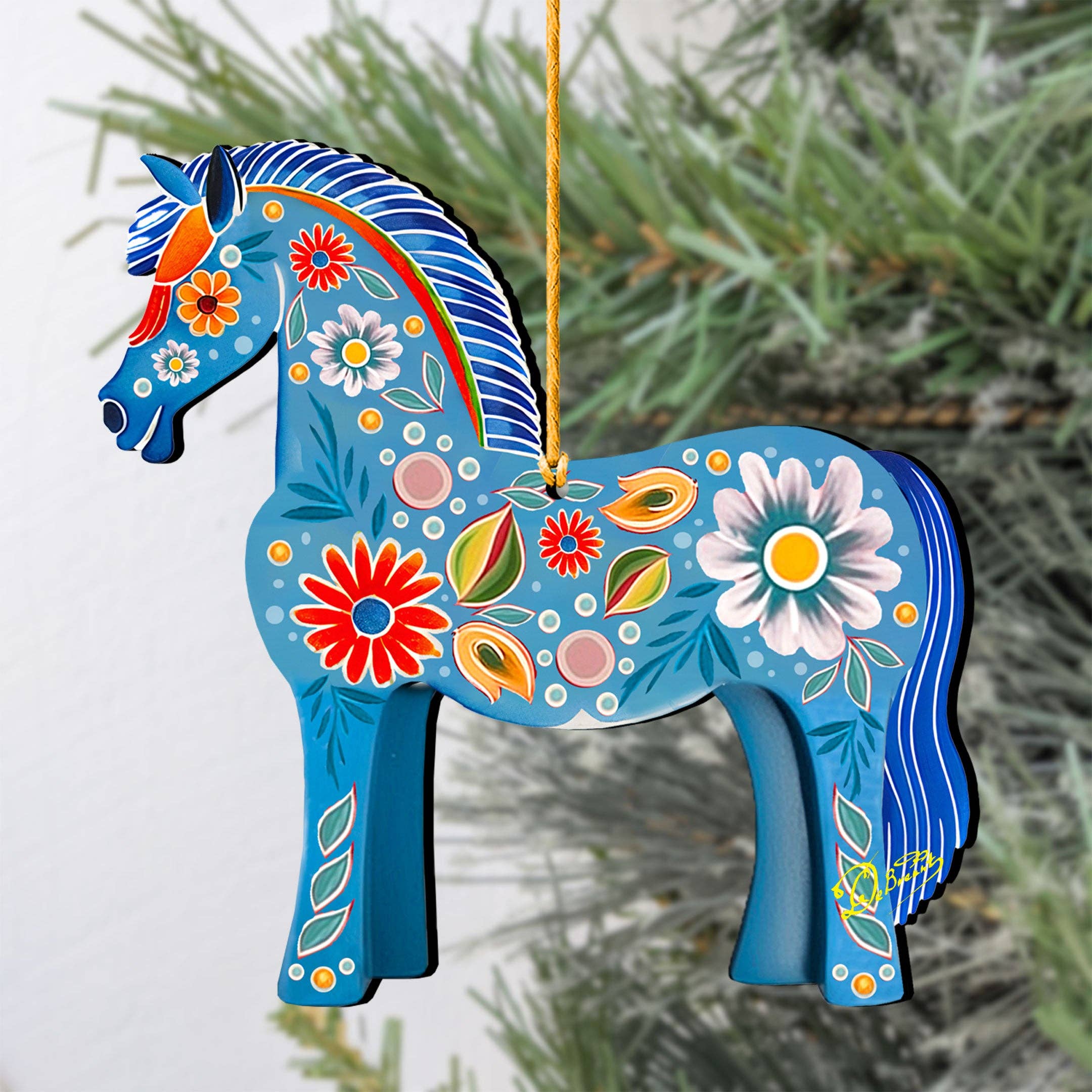 The Danish Horse Decorative Wood Christmas Tree Ornament by G Debrekht (5.5"x5") features a blue horse with colorful floral patterns, hanging on a yellow string in front of a green pine branch.