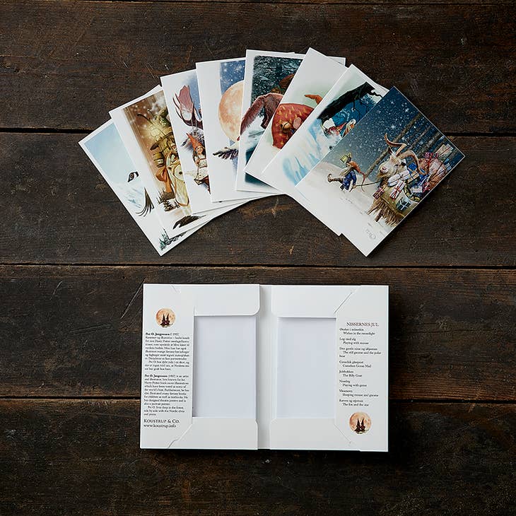 The Card Pack: Nissernes Jul Christmas with Nordic Elves & Pixies by Koustrupco, featuring 8 different illustrated cards with envelopes, is arranged in a fan above an open cardholder on a dark wooden surface.