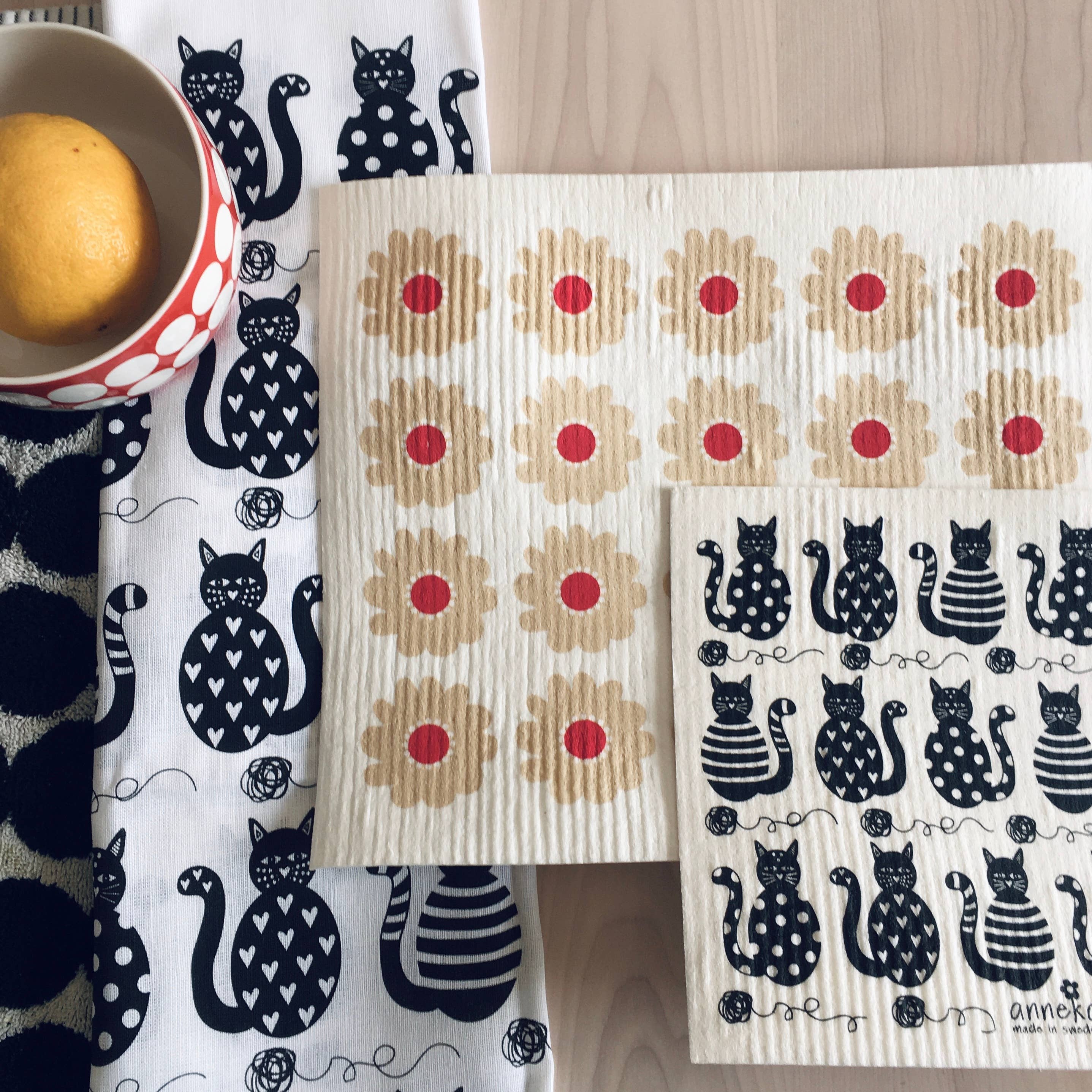 A lemon in a red and white polka-dot cup sits beside two Cats Swedish Dishcloths by Anneko Design Sweden, featuring black cat patterns, and two rectangular cloths with red and beige floral prints on a light wooden surface.