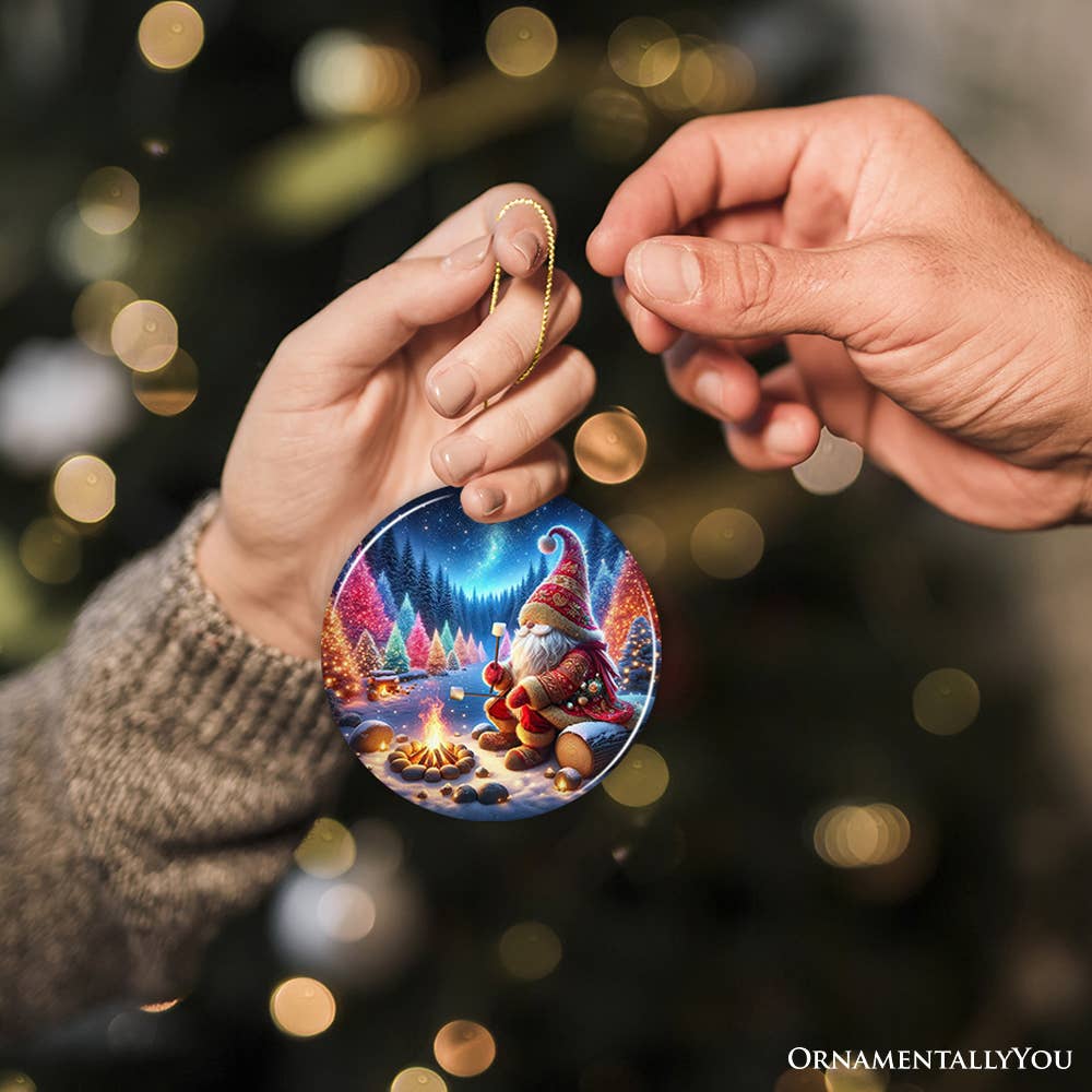 Two hands hold the Majestic Winter World Gnome Camper Ceramic Ornament, showcasing a winter gnome by a campfire, with a blurred Christmas tree in the background.