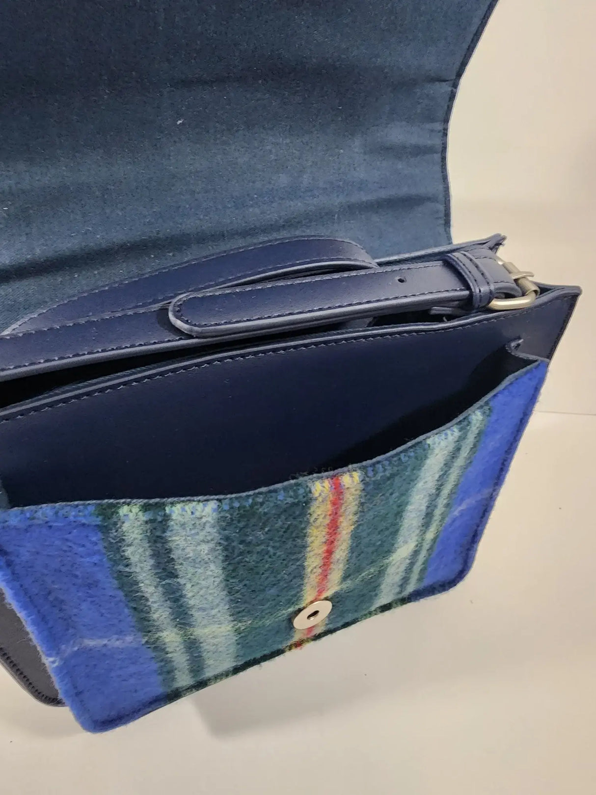 The Nova Scotia Patsy Handbag is a blue leather bag featuring an open flap with an inner handle, and an exterior pocket adorned with multicolored provincial tartan fabric and a button closure.