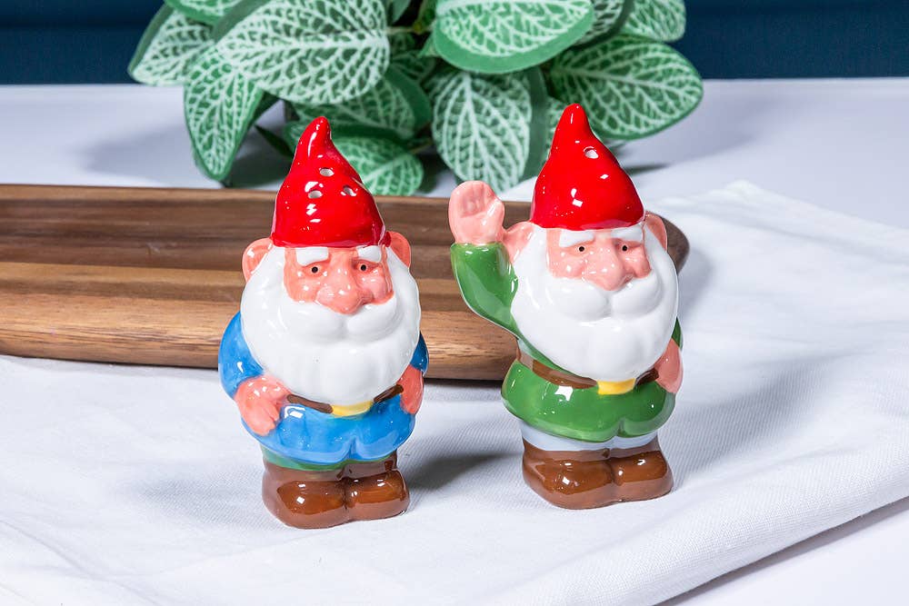 Shakers: Garden Gnomes - Salt & Pepper Shakers feature two ceramic gnomes with red hats, white beards, and colorful outfits on a white cloth; one has its hand raised. A plant and wooden board are visible in the background.