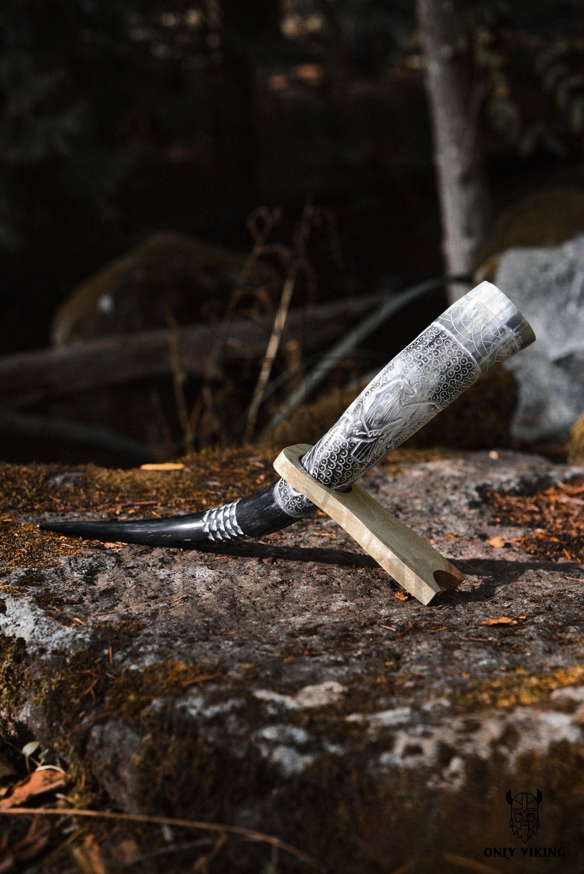 The Horn: Raven Viking Drinking Horn w/ Metal Stand (14"-18") featuring intricate raven engravings is displayed on a mossy rock in an outdoor setting.