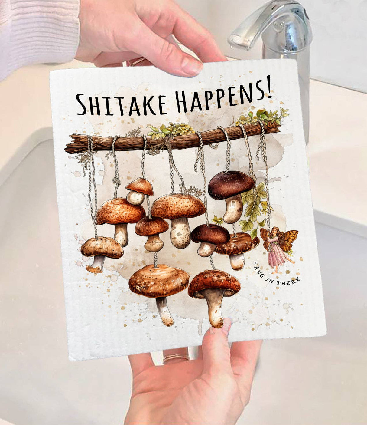 A person holding a compostable Dish Cloth: Shitake Happens! Hang in There - Mushrooms and Fairy, featuring illustrated shiitake mushrooms hanging from strings and the playful text Shitake Happens! Hang in there, near a sink.