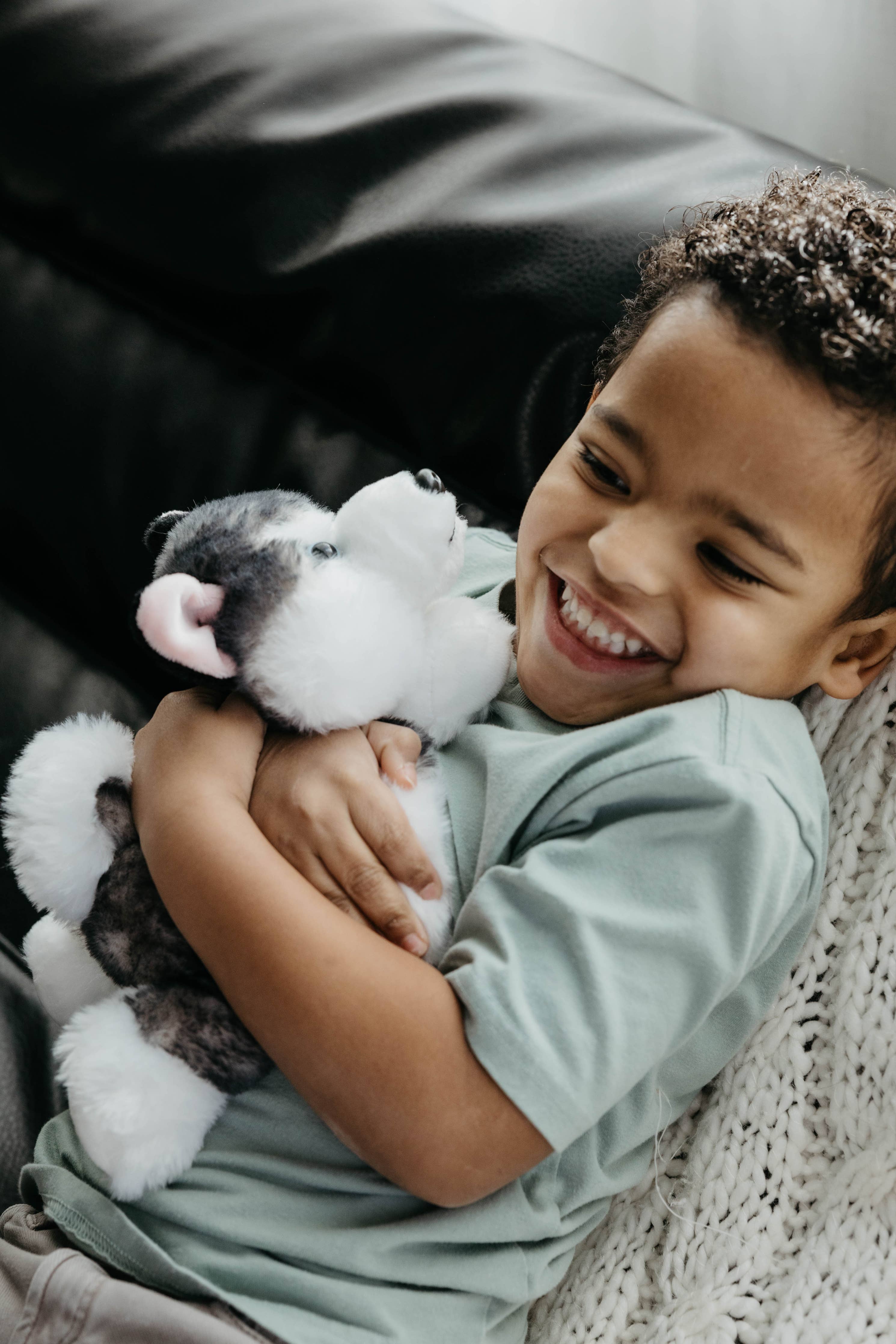 A young child sits on a couch, smiling and hugging the 9 Plush Husky Dog Stuffed Animal while wrapped in a knitted blanket.