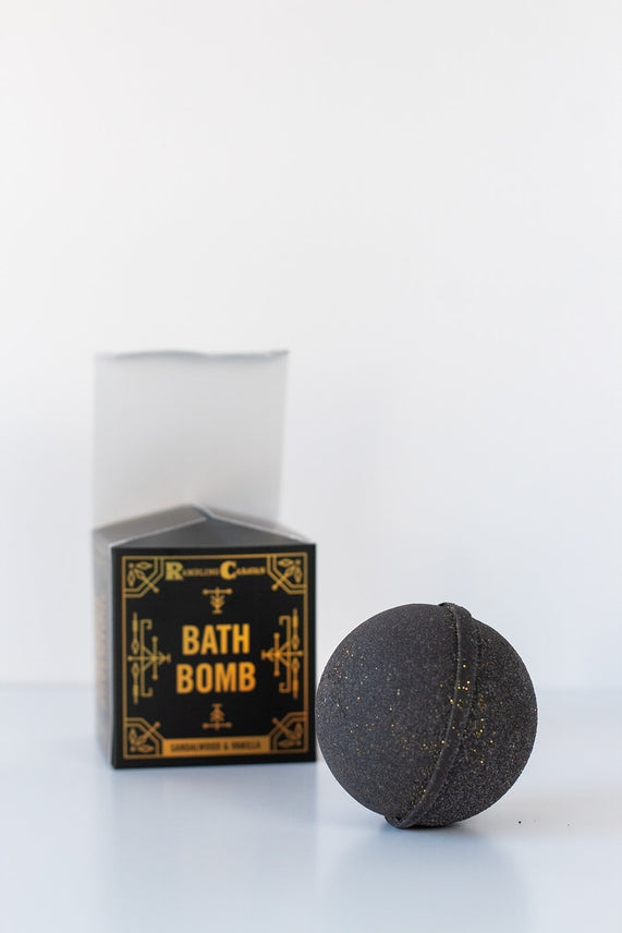 A black Sandalwood & Vanilla Bath Bomb with gold specks sits before its black and gold Bath Bomb packaging, emphasizing its handmade quality and nourishing ingredients.