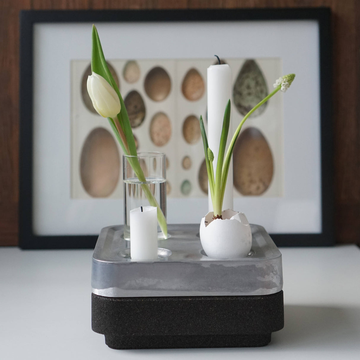 The Candle Holder: Mini Vases for Stumpastaken Candleholder 2-pack features white tulips and plants in eggshells, arranged with a candle on layered surfaces and framed egg art, making a charming table decoration.