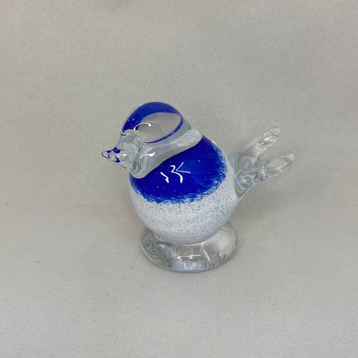 The White & Blue Tipu Blown Glass Bird by Bianco Blue, designed by Tarmo Maaronen, features a blue head and upper body with clear wings and detailed facial features. It stands elegantly on a clear base against a simple background.