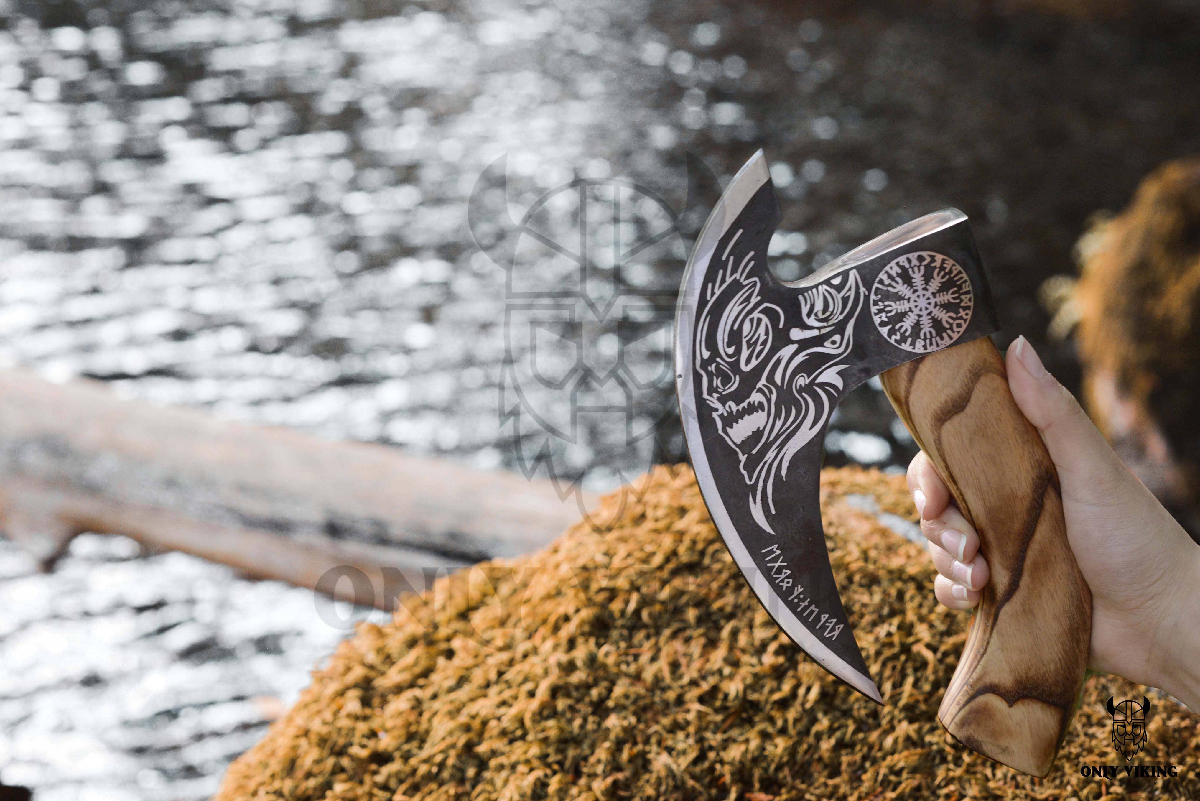 By a body of water, someone holds a stylish Pizza Cutter: Axe & Leather Axe Sheath, featuring an ornately designed handle and engraved stainless steel blade.