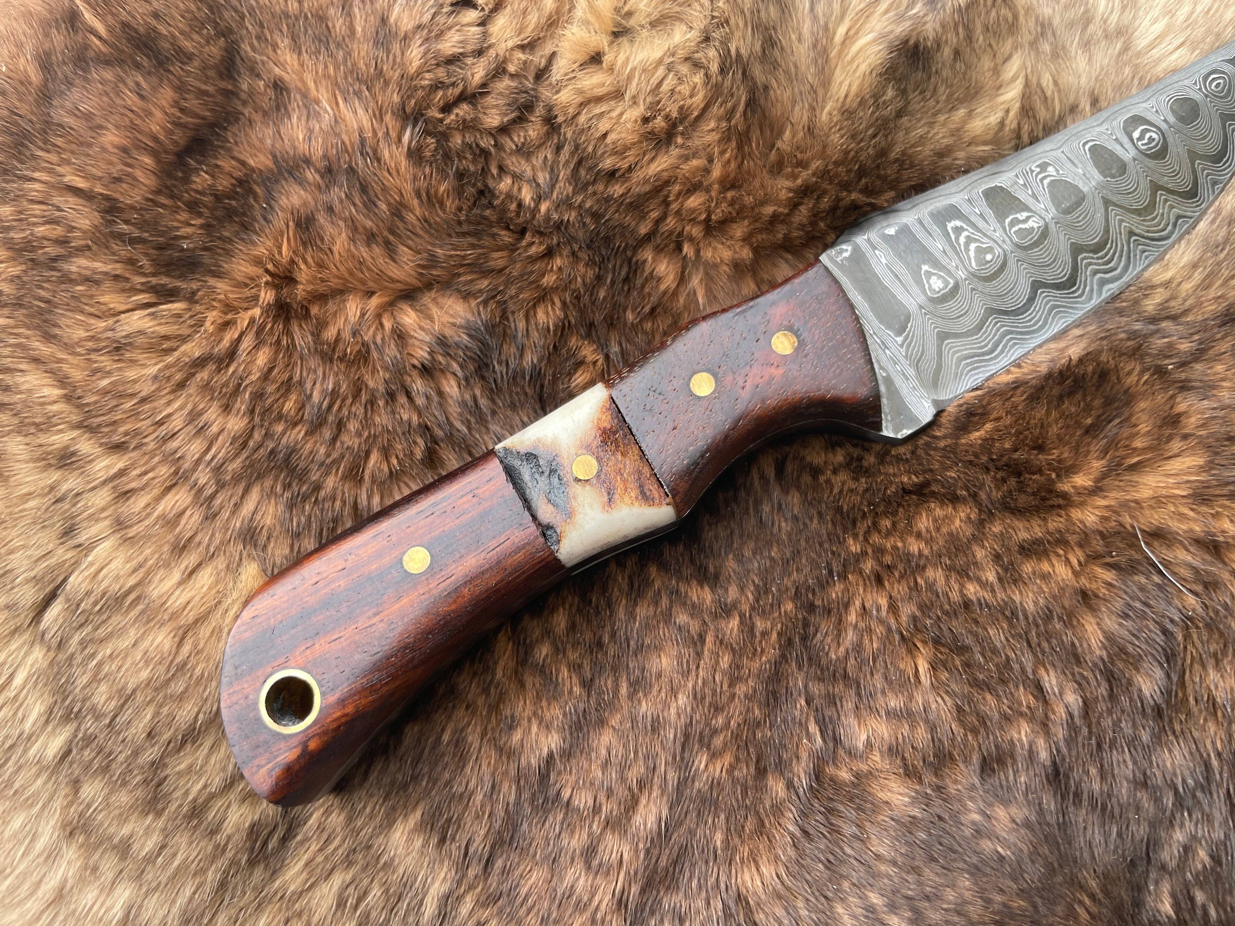 The Knife: Damascus Steel Skinner Knife with Rosewood & Stag Grip features a Damascus steel blade and a handle made of rosewood and stag, enhanced with metal and bone accents, shown resting on brown fur.