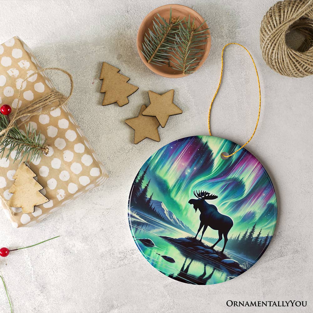 The "Majestic Moose Under Northern Lights Christmas Tree Ornament" is beautifully set in a festive scene featuring nature-inspired holiday decor. This includes wrapped gifts, star-shaped cookies, pine branches, and string for hanging.