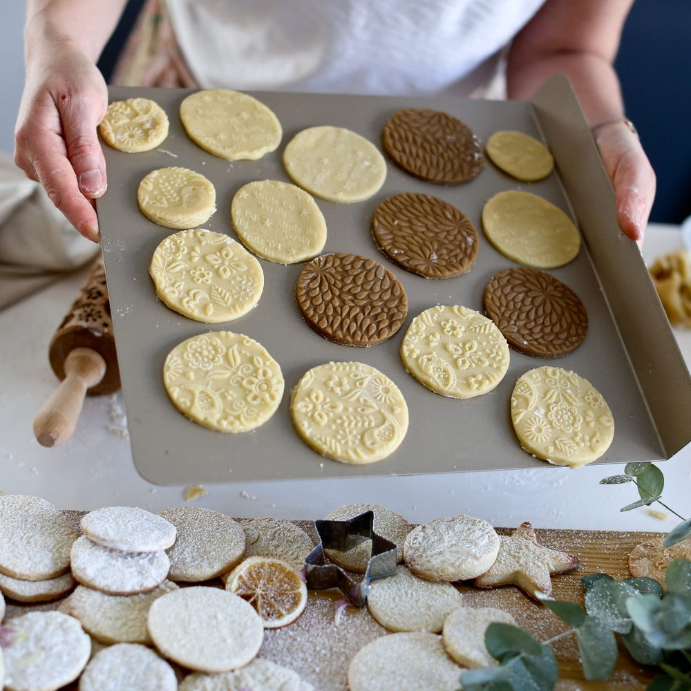 A person holds a baking tray adorned with cookies featuring Scandinavian-inspired patterns, ready for the oven. The dough was rolled using the Reindeer Birds & Trees Embossing Rolling Pin and cookie cutters are scattered on the table.