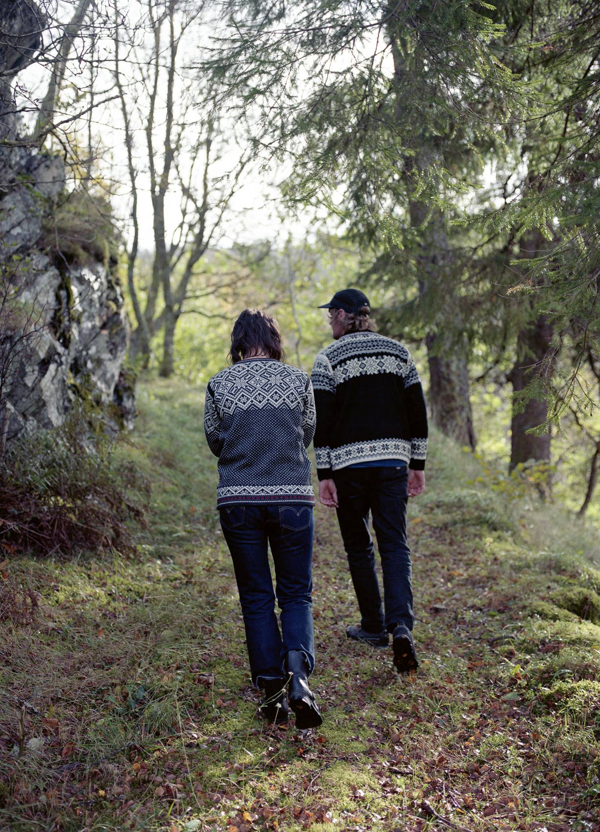 Two people wearing Stavanger Norwegian Wool Cardigans in black walk up a grassy, wooded path surrounded by trees and rocks on a cloudy day.