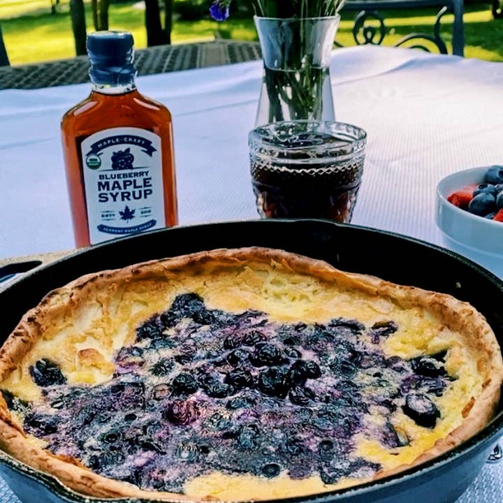 A blueberry pancake in a skillet sits on a table, accompanied by antioxidant-rich Summer Favorites Blueberry Maple Syrup (6.8 oz), a glass of dark liquid, and a bowl of fruit.