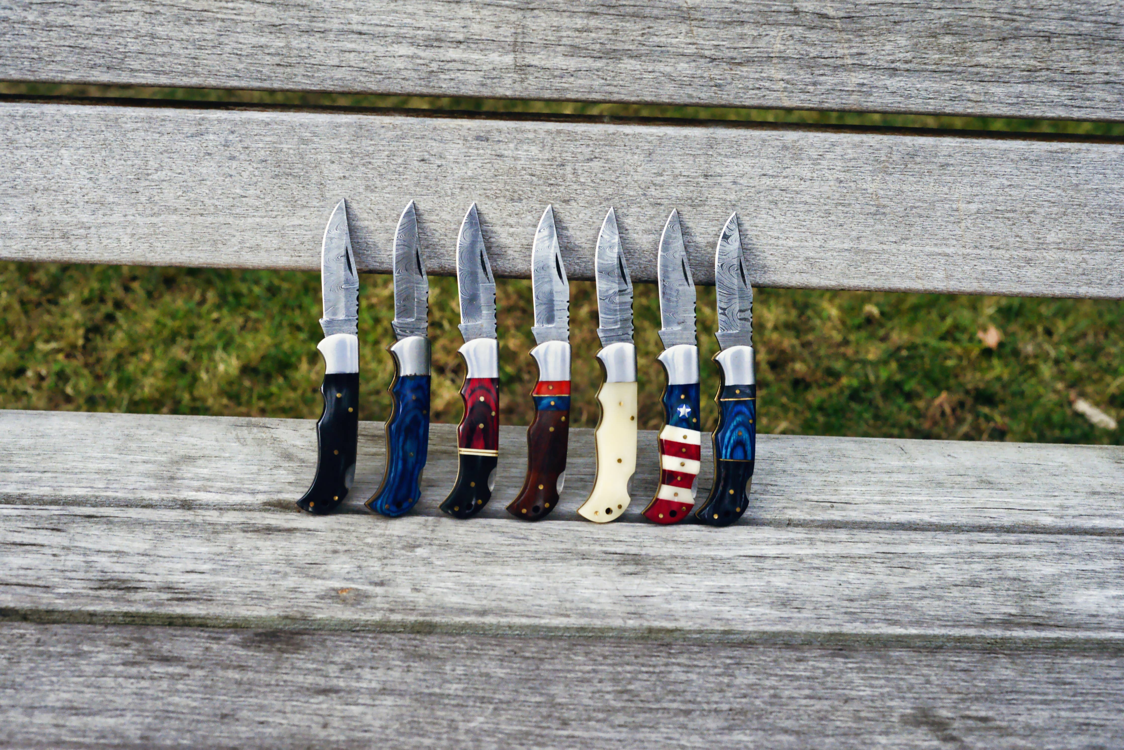 Seven folding knives, including the striking Camel Bone Handle Genuine Damascus Steel Folding Knife, are displayed on a wooden bench outdoors.