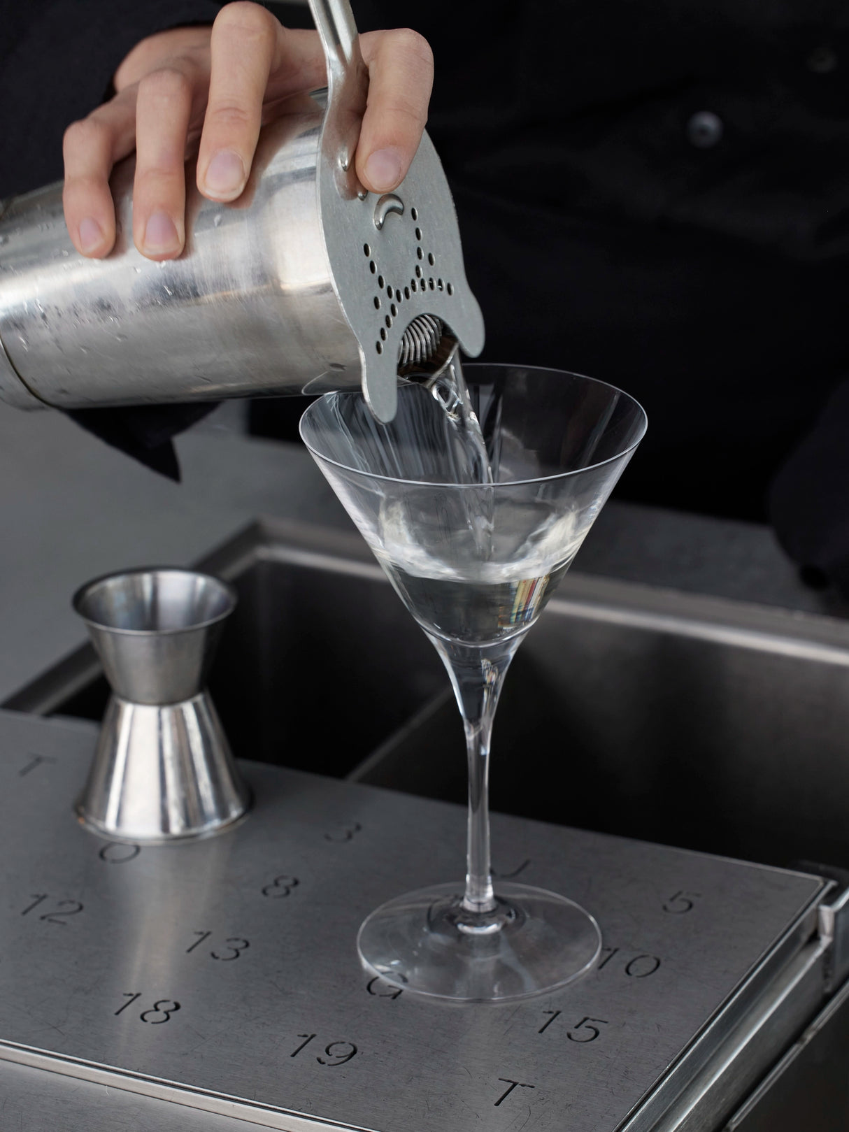 A bartender gracefully pours a clear liquid from a shaker into an Orrefors: More Martini Glass 6.4oz (2-Pack). A numbered jigger sits nearby on the metal counter, ready for the next creation.