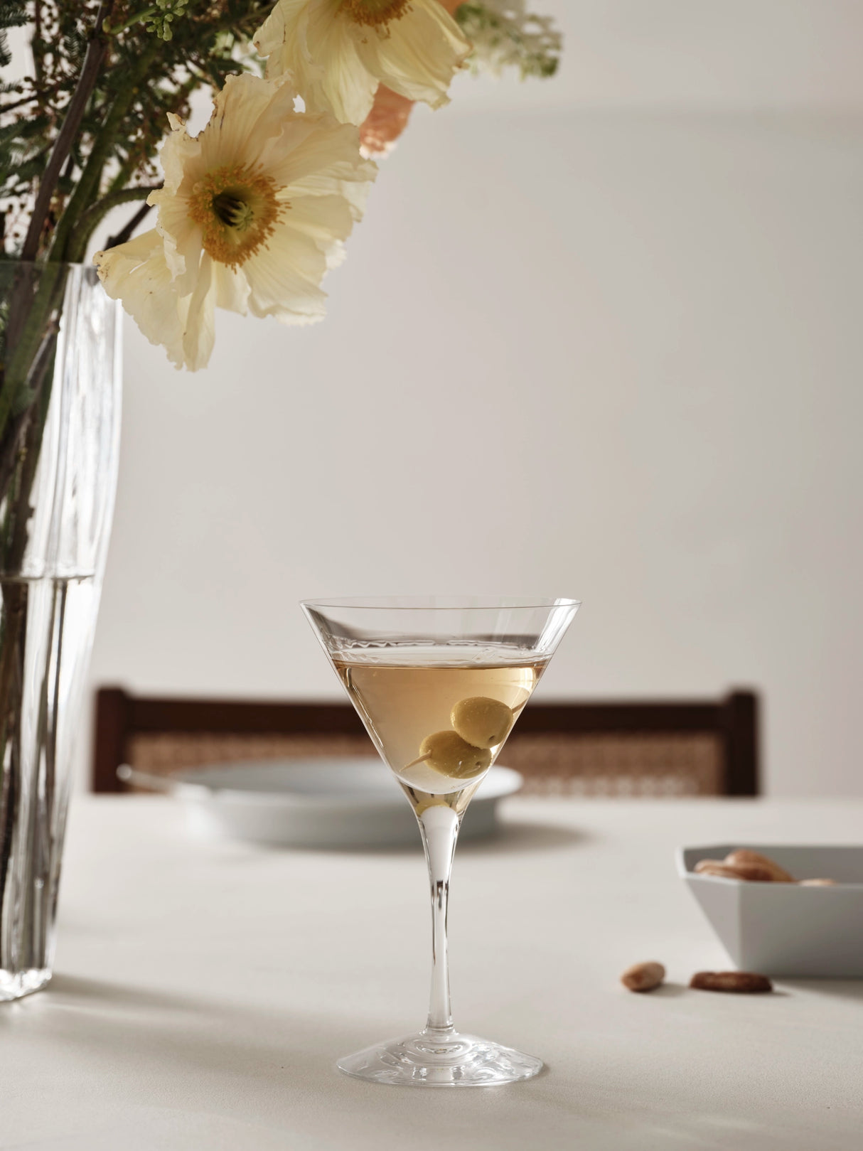 An Orrefors: More Martini Glass 6.4oz from the 2-pack, filled with olives, sits beside a large flower vase and a bowl of nuts, showcasing Orrefors elegance.