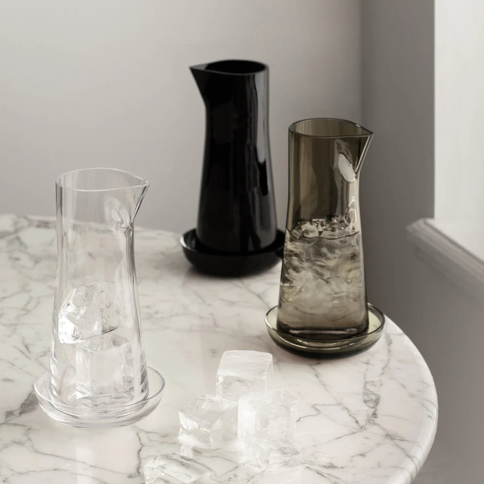 An Orrefors Informal Carafe with Bowl in olive glass sits on a round marble table, with large ice cubes scattered nearby and inside the carafe.