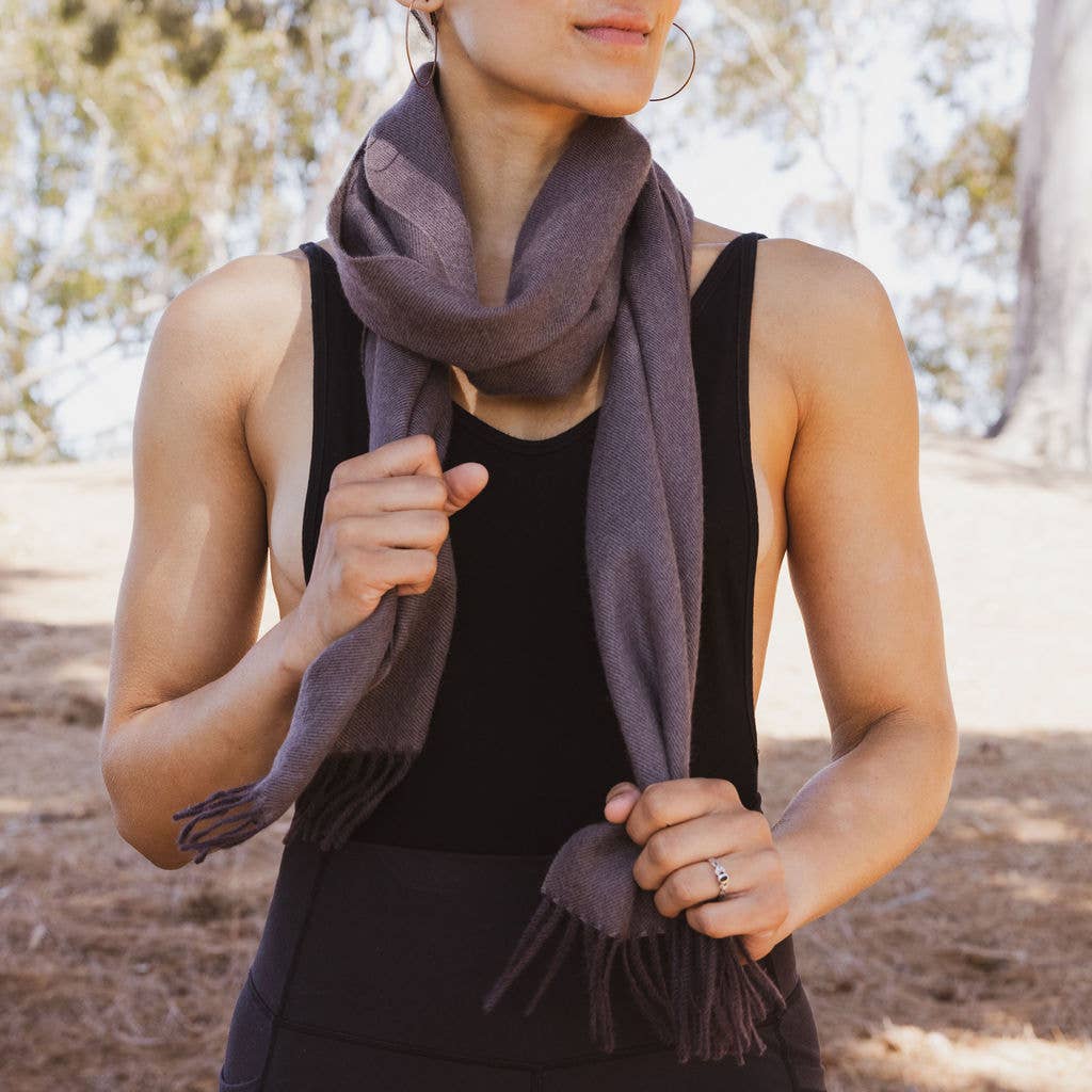 A person in a black sleeveless top is wearing the Truffle 100% Baby Alpaca Scarf, standing outdoors with trees in the background. The luxury warmth of this finest fiber offers both style and comfort amidst nature's embrace.