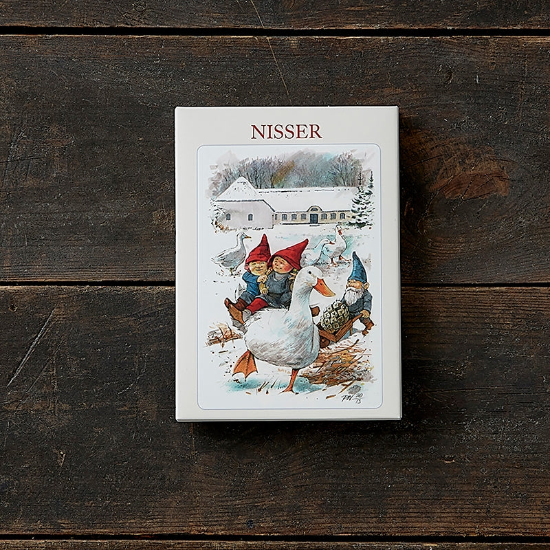 Box of holiday cards titled "Card Pack: Nisser" by Koustrupco, featuring artwork by Peter Nielsen of elves riding geese in front of a snowy house, displayed on a wooden surface.