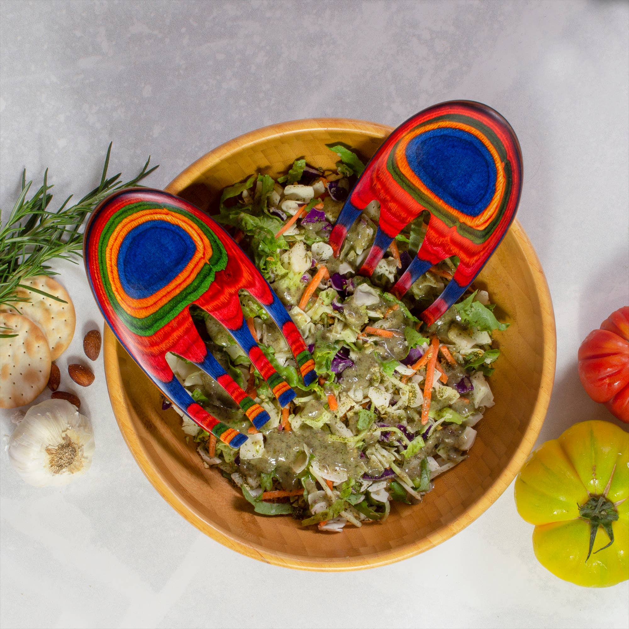 A wooden bowl of salad is served with Utensil: Colorful Birch Salad Hands, surrounded by garlic, crackers, rosemary, and two heirloom tomatoes on a light countertop.