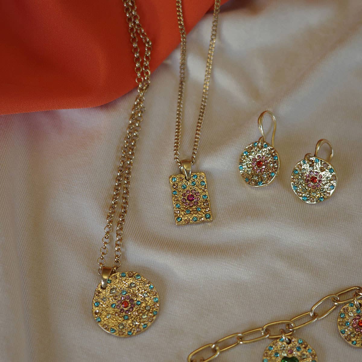 The A&C Oslo Vibrance Gold Coin Earrings are displayed with gold jewelry, including a necklace, layered pendants, and a bracelet adorned with multicolored gemstones on white fabric and an orange cloth background.