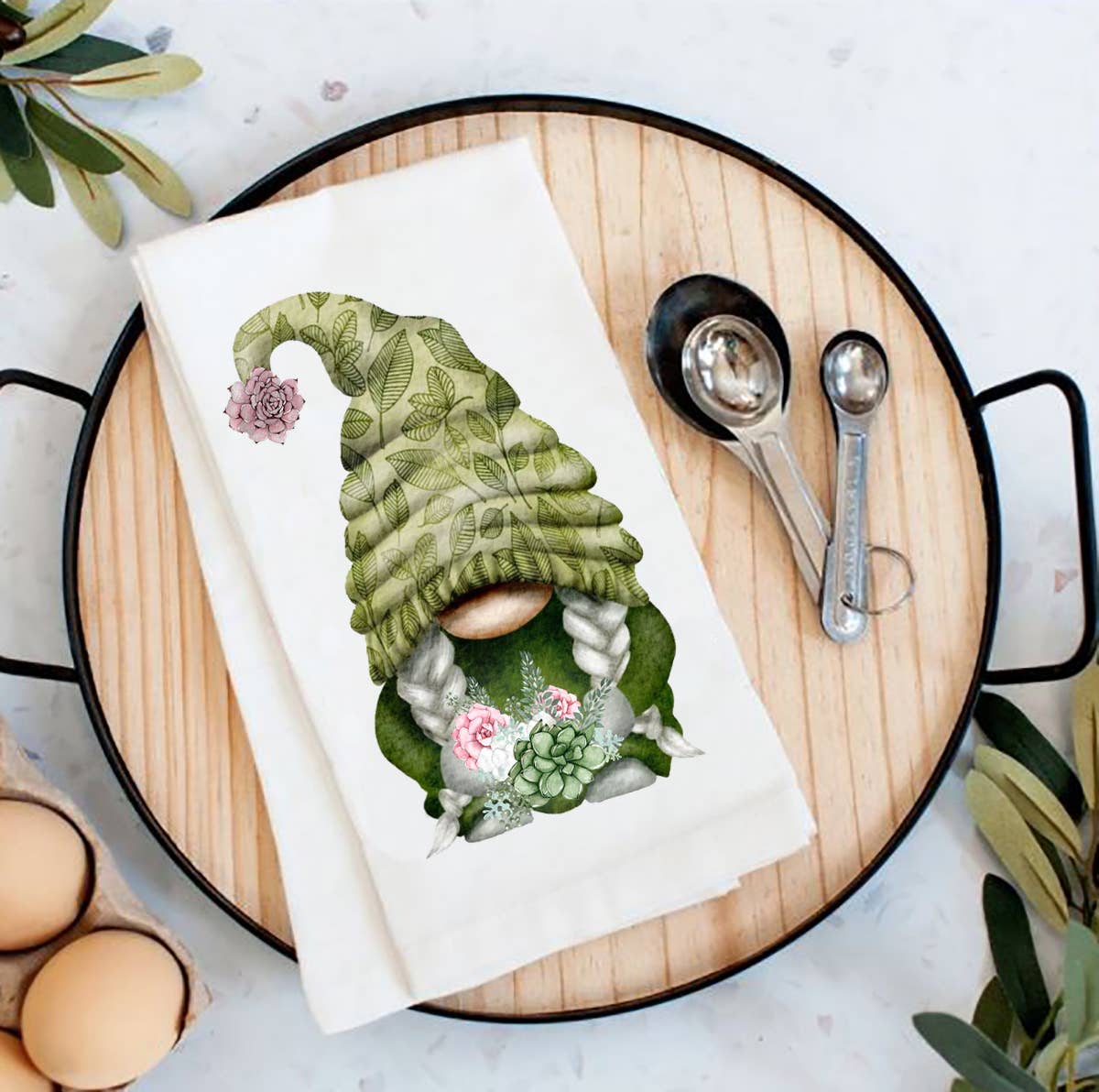 A wooden tray features a charming Succulent Desert Green Gnome Flour Sack Tea Towel, ideal for farmhouse decor. It includes measuring spoons on the side, eggs in a carton, and plant leaves tucked in the corners.