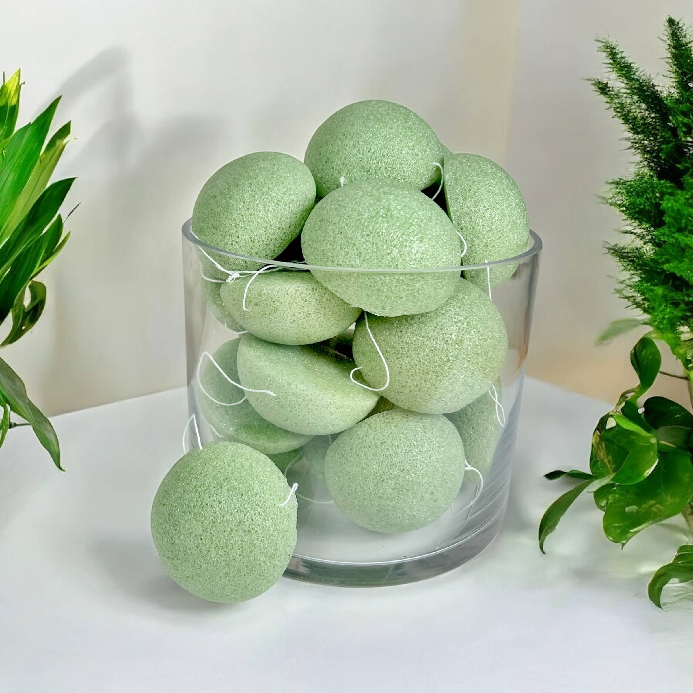 A clear glass container holds seafoam green, oval-shaped Natural Konjac Facial Cleansing Sponges, with a few of these sponges and green plants arranged nearby for gentle exfoliation.