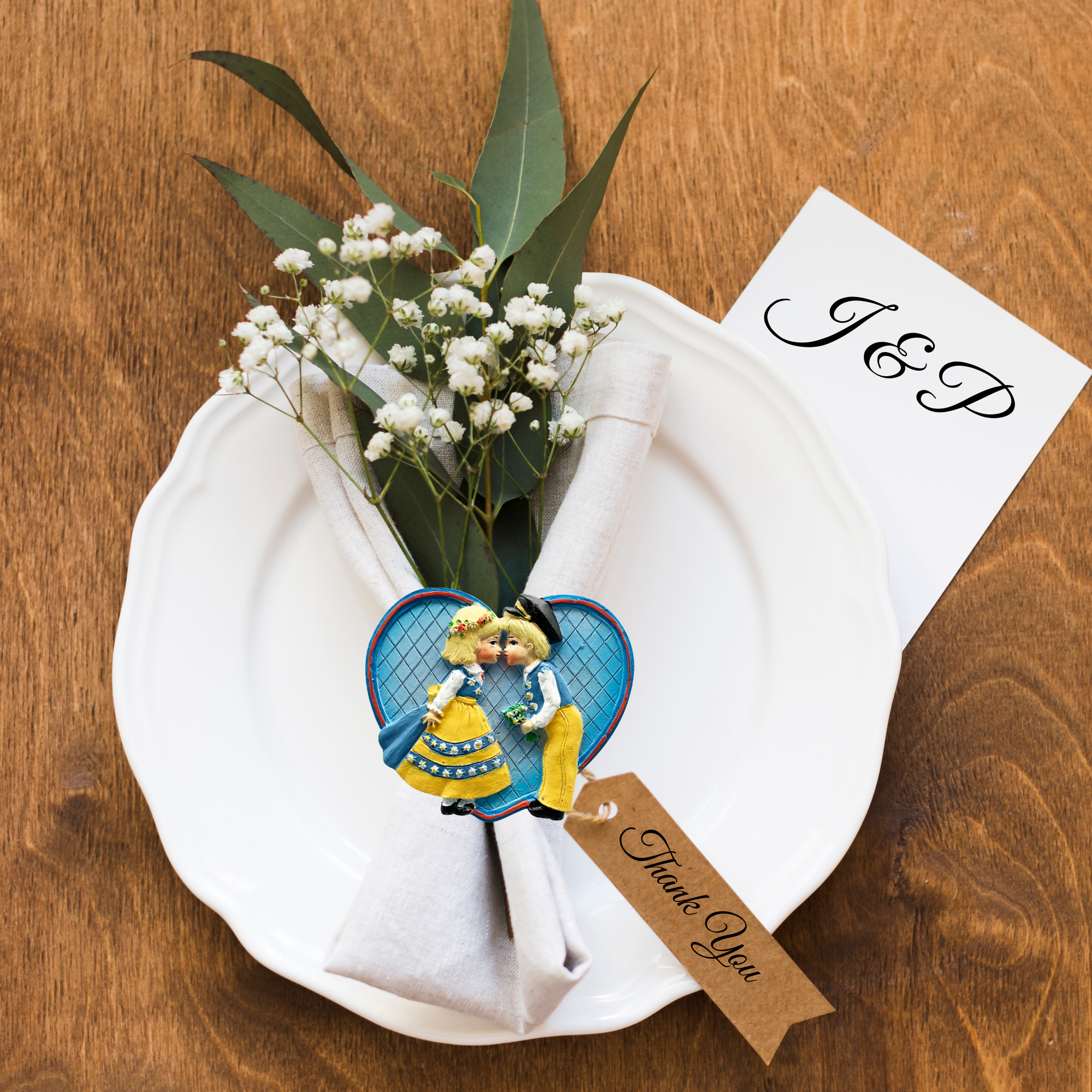 A decorative napkin with a small bouquet, thank you tag, and novelty gift heart sits on a white plate. Nearby on the wooden table rests a card with J & P and the Magnet: Swedish Kissing Couple Party Favor Kitchen Magnet, adding charm to the setting.